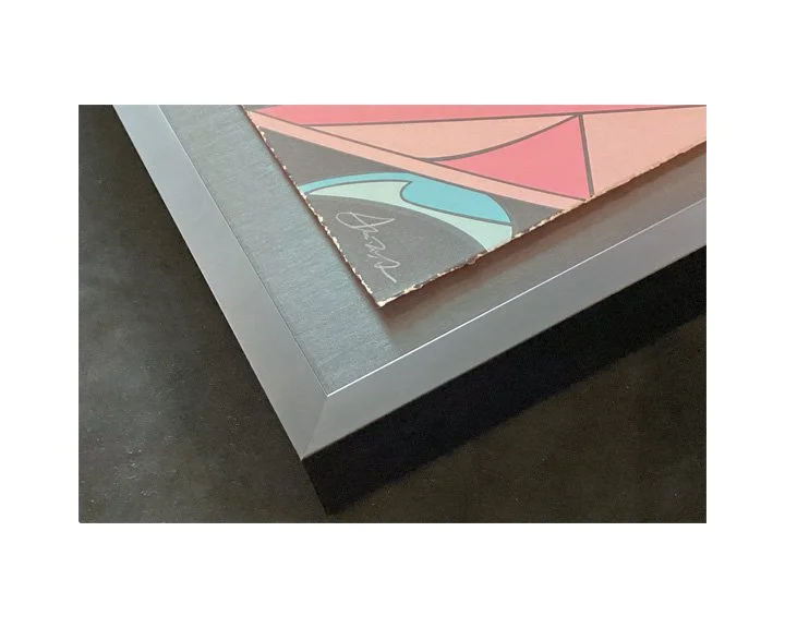 Close-up of corner of framed abstract artwork with geometric shapes and signature, on a dark surface.
