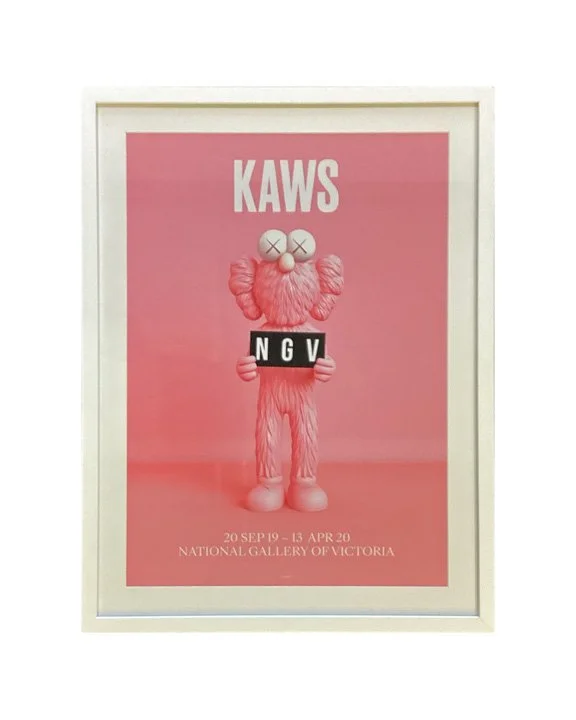 Pink poster for an art exhibition featuring a pink furry creature with cross eyes, holding a black sign with white letters 'NGV', titled 'KAWS' with exhibition dates from September 20, 2019, to April 13, 2020, at the National Gallery of Victoria.