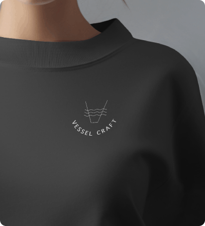 Close-up of a black sweatshirt with a small white logo that reads "VESSEL CRAFT" and features a simple line drawing of a boat.