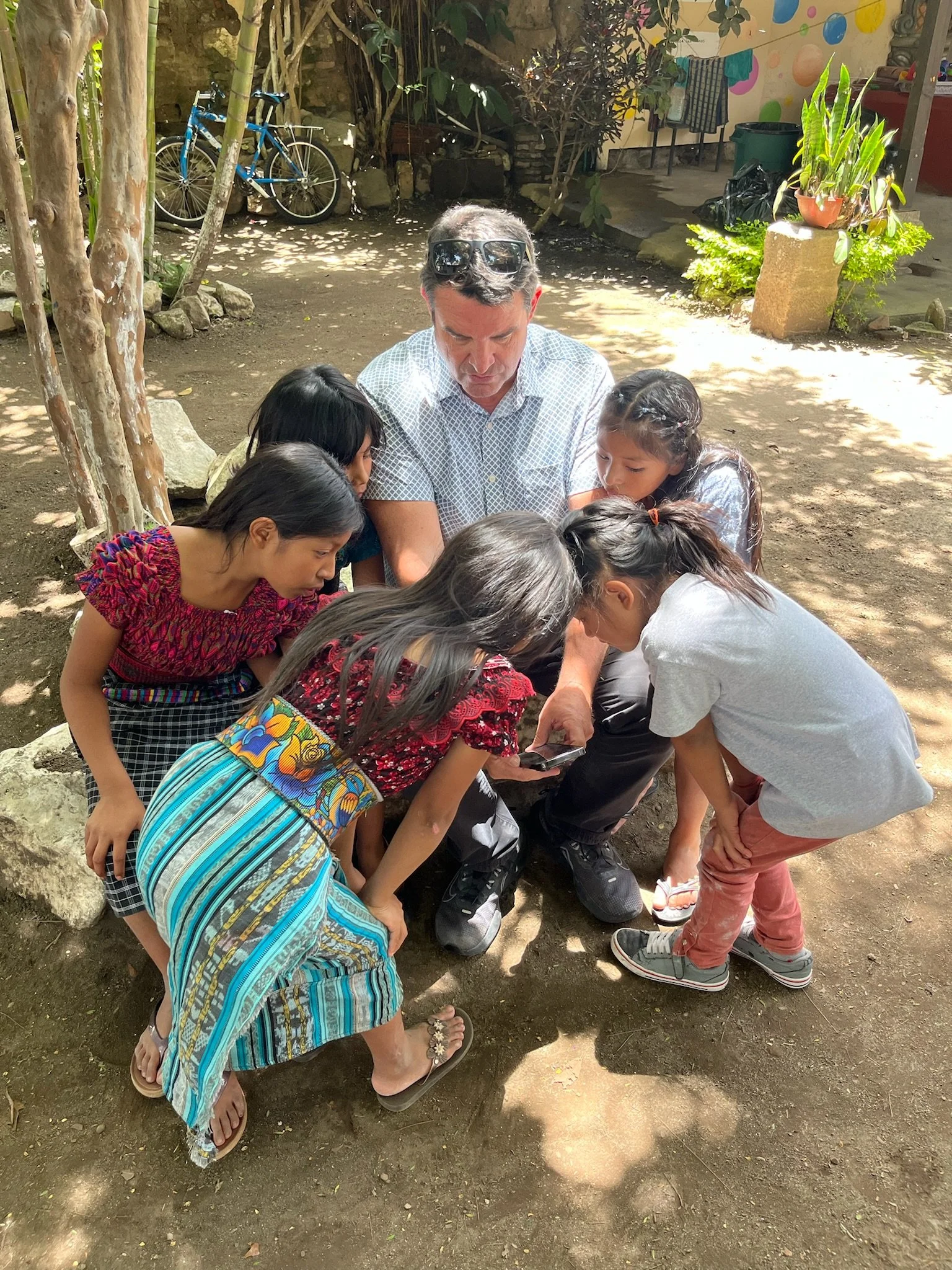 Man with sunglasses on his head and five young girls gathered around him, all looking at a phone held by one of the girls, outdoors in a shaded area.
