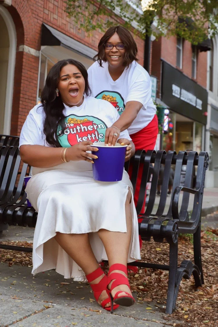Southern Kettle Co. founder Kaneisha Dotson and co-founder Desiree McMurry-Ellis enjoying popcorn together on a park bench.