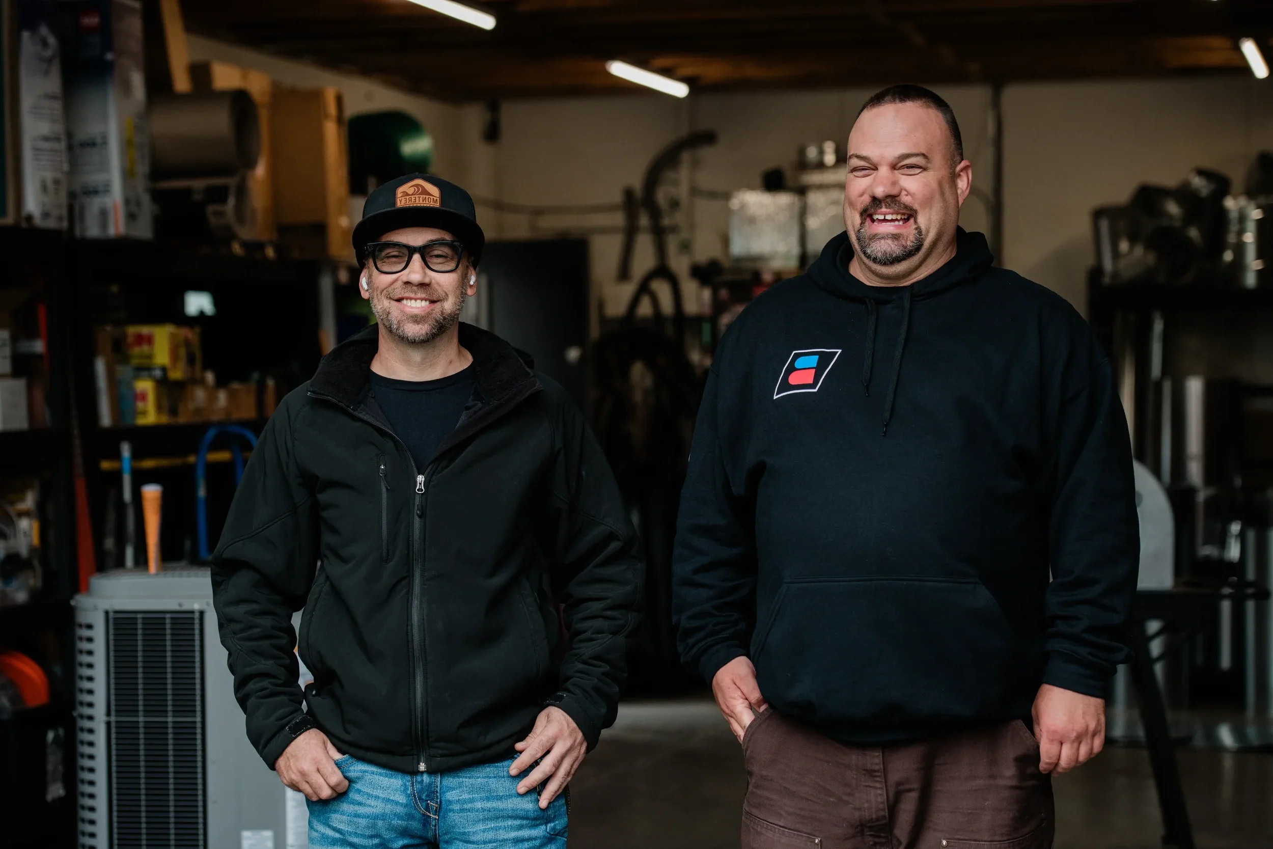 Two men standing in a cluttered workshop. The man on the left wears glasses, a black jacket, and a cap, while the man on the right wears a black hoodie with a logo and brown pants.