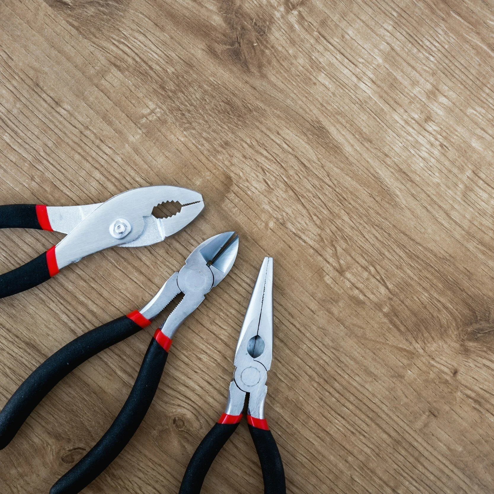 Three different types of pliers with black and red handles on a wooden surface.