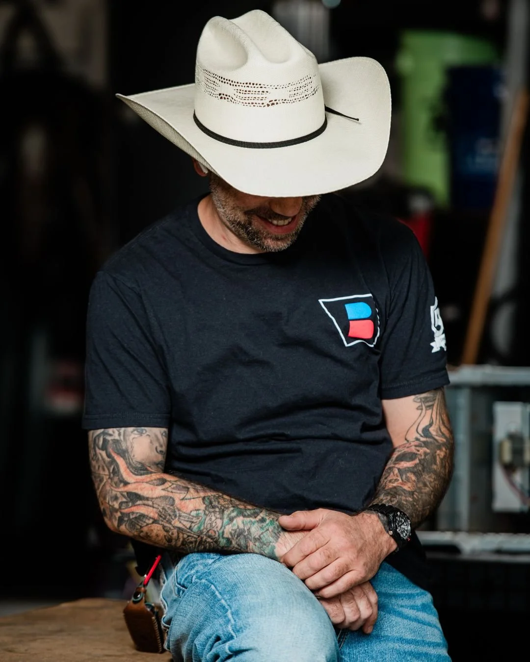 A man wearing a white wide-brimmed cowboy hat, a black t-shirt with a red and blue logo, and jeans, sitting with his hands clasped and looking down, smiling, with tattoos visible on his arms.