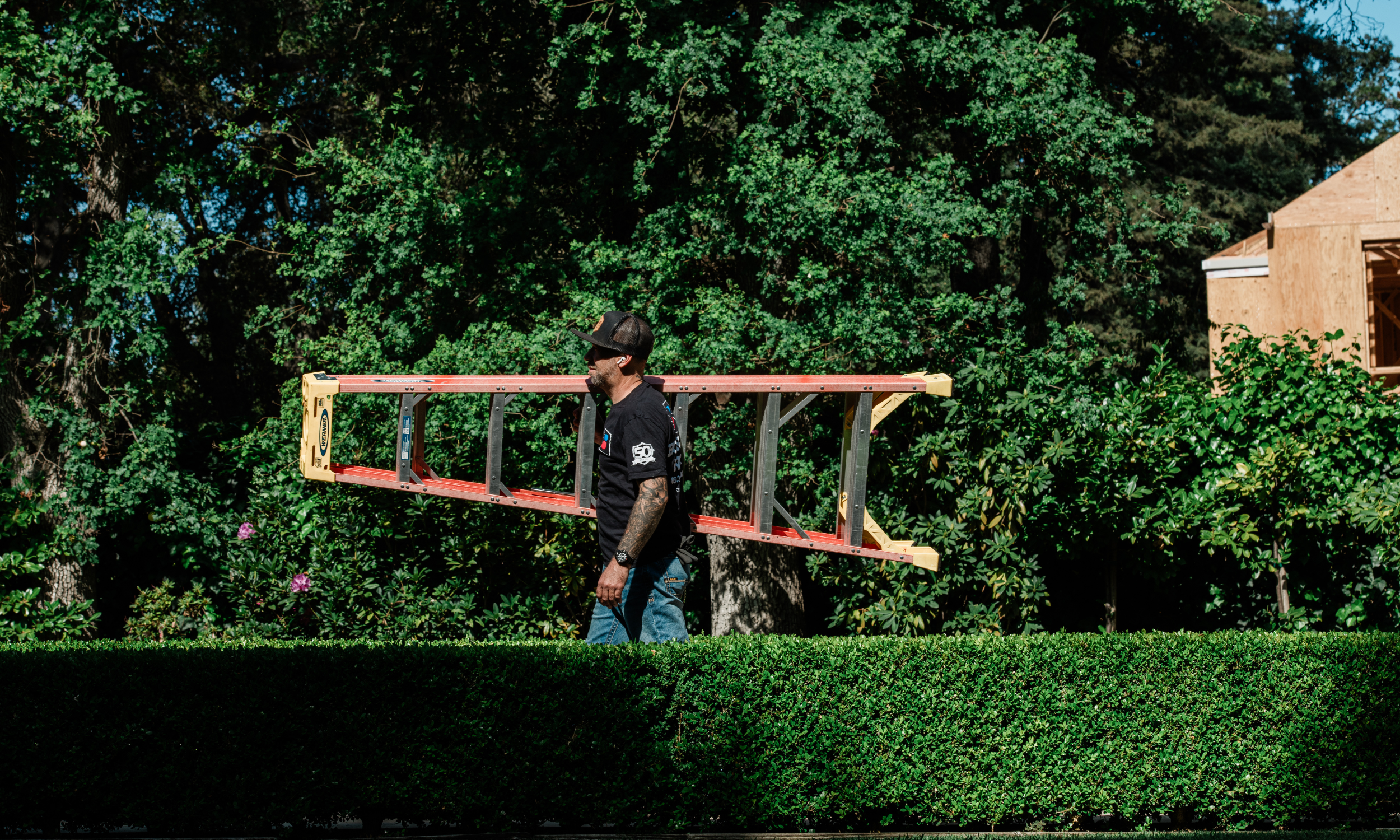 Professional installer John carrying a Werner fiberglass extension ladder across a landscaped lawn for a project in Atherton.