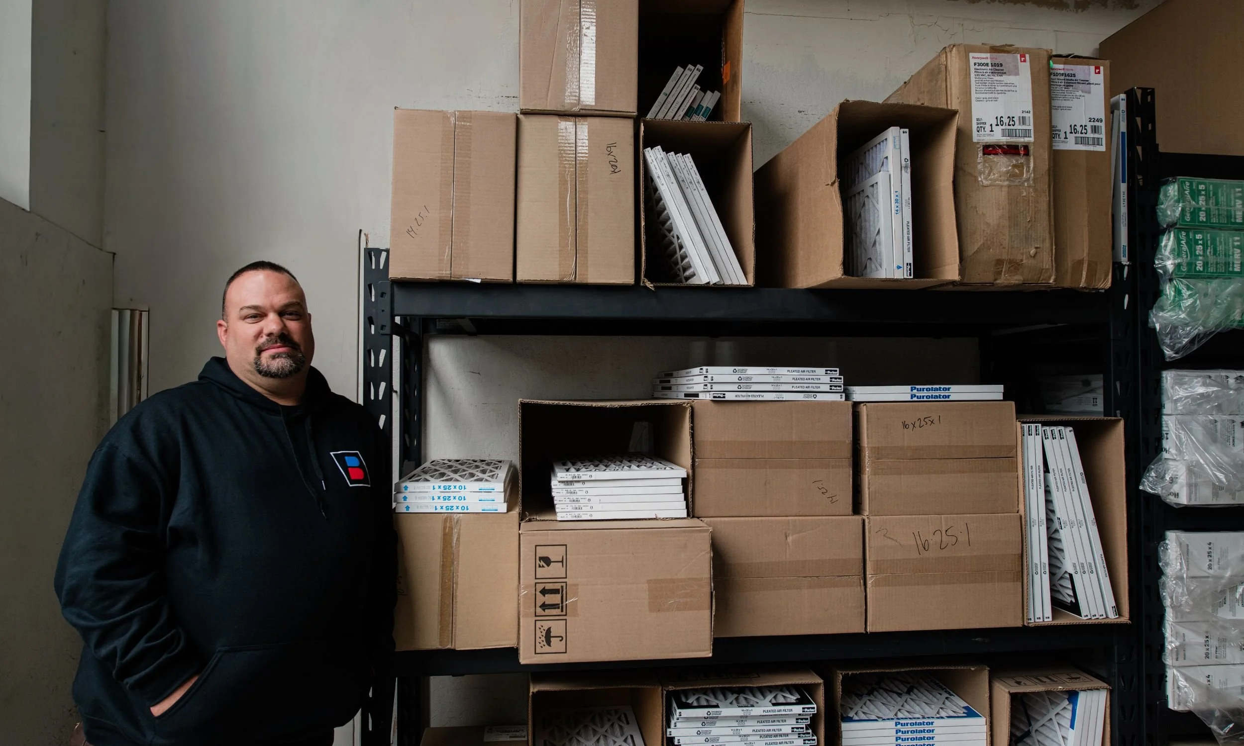A man with a goatee and short hair standing in front of shelves filled with various boxes and filters in a warehouse or storage room.