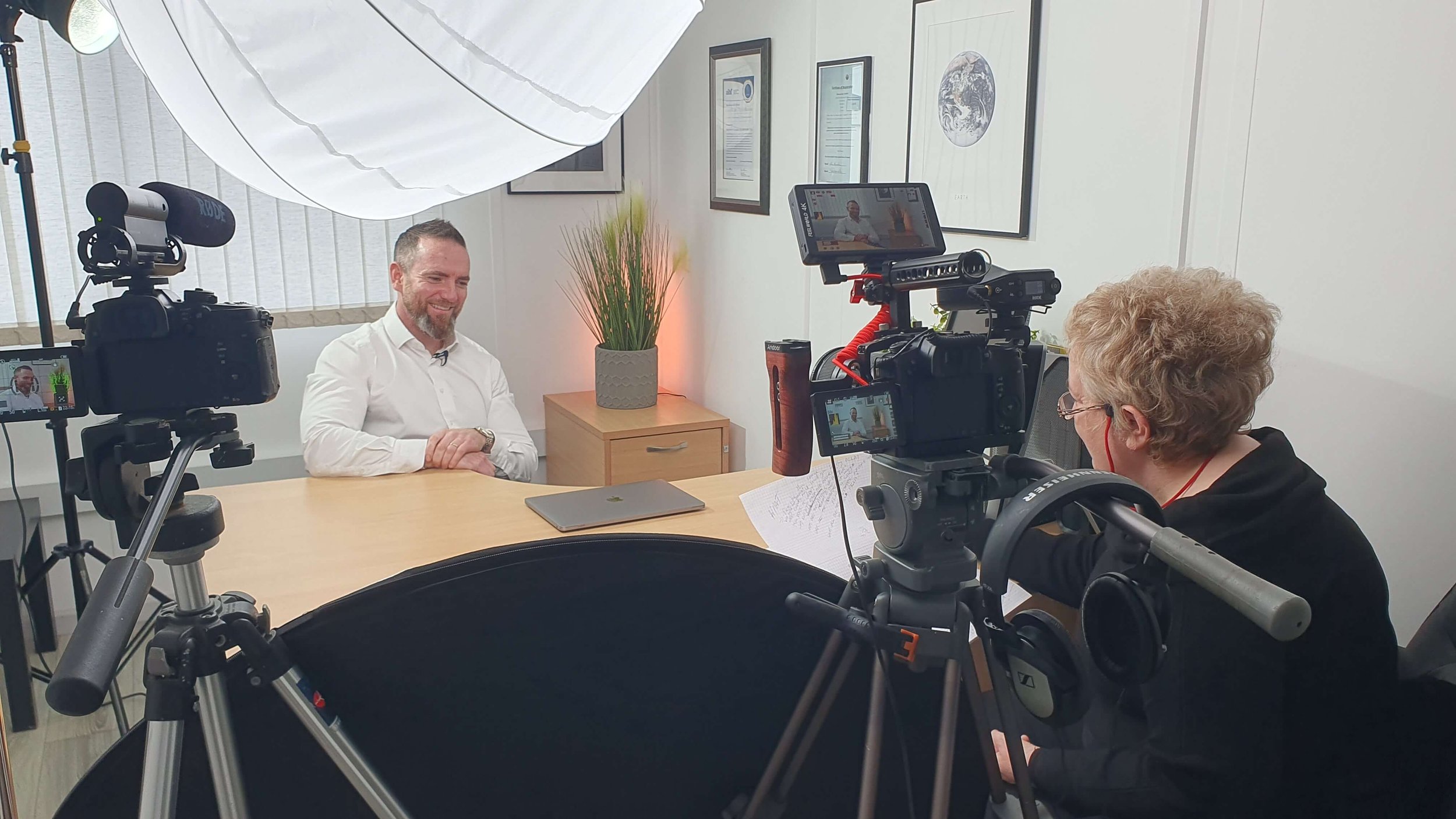 A man with a beard and white shirt being interviewed in an office about a camera crew filming the session. The setting is a professional interview with lighting, cameras, and documents on the table.