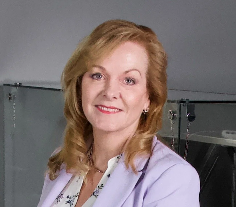 A woman with shoulder-length red hair and a light purple blazer smiling at the camera, in an indoor setting with a gray background.