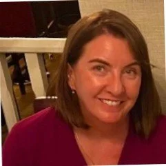 A woman with shoulder-length brown hair smiling, wearing a dark pink or purple top, indoor background.