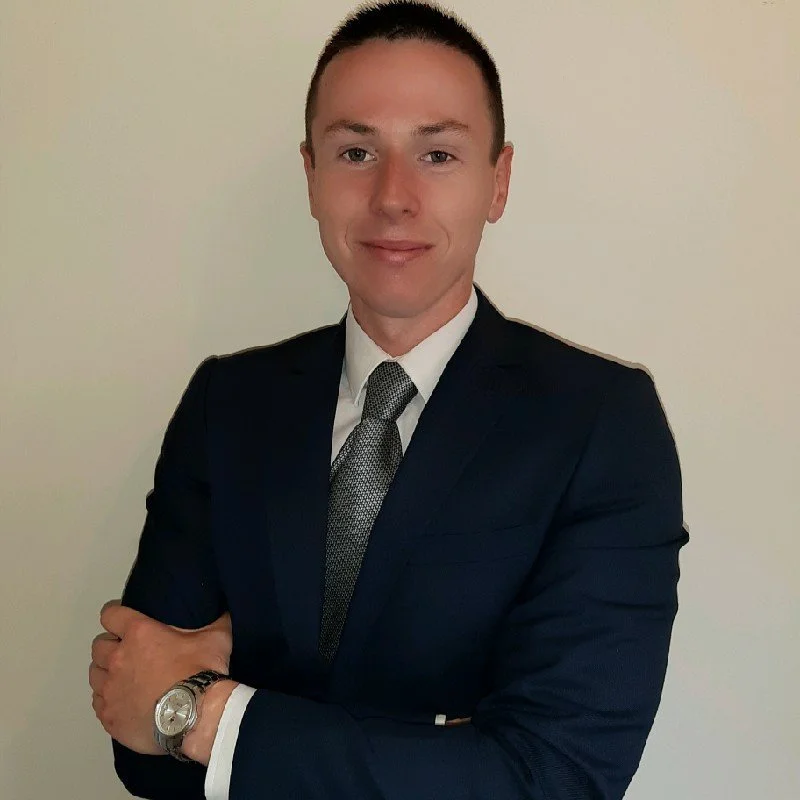 A man in a dark suit, white shirt, and patterned tie standing with arms crossed against a plain background.
