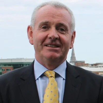 A middle-aged man in a suit and yellow tie standing outdoors with the cityscape and a building with a spire in the background.