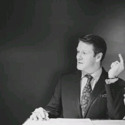 A man in a suit sitting at a desk with his right hand raised, looking to the side, in black and white.