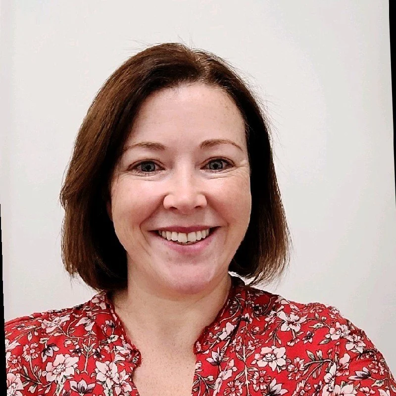 A woman with shoulder-length brown hair, smiling, wearing a red floral-patterned blouse, standing in front of a plain white wall.