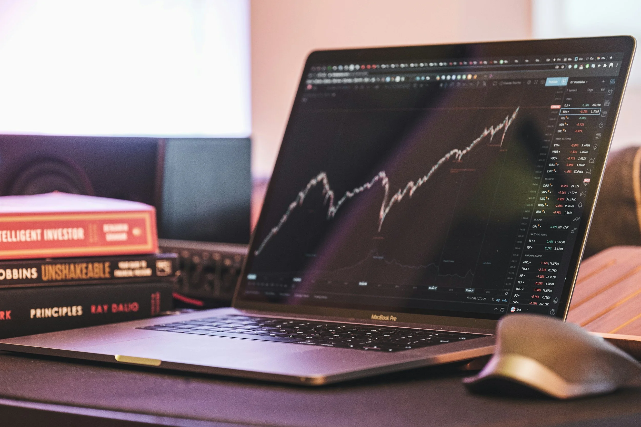 A MacBook Pro laptop displaying a stock market trading chart in front of a computer monitor and a mouse. There are books about investing and finance on the desk.