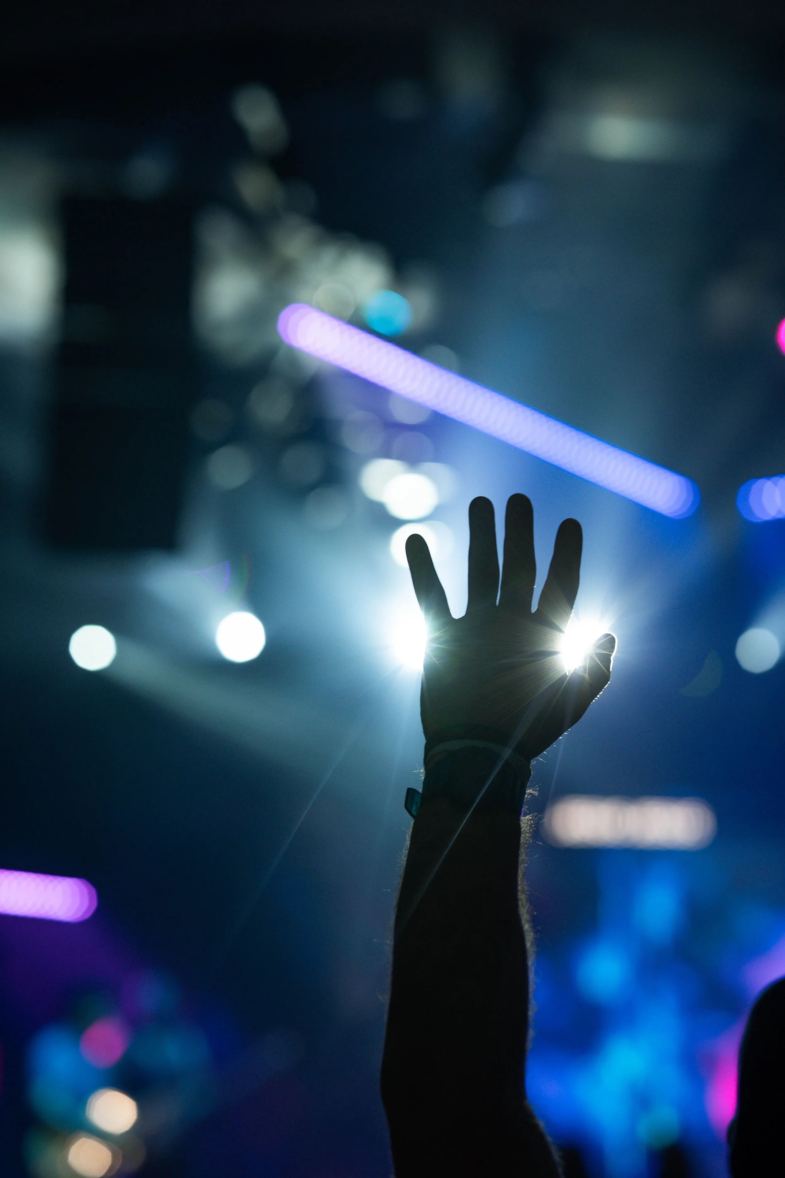 A person raising their hand with fingers spread, illuminated by bright lights in a dark concert or nightclub setting with colorful lights and bokeh effects in the background.