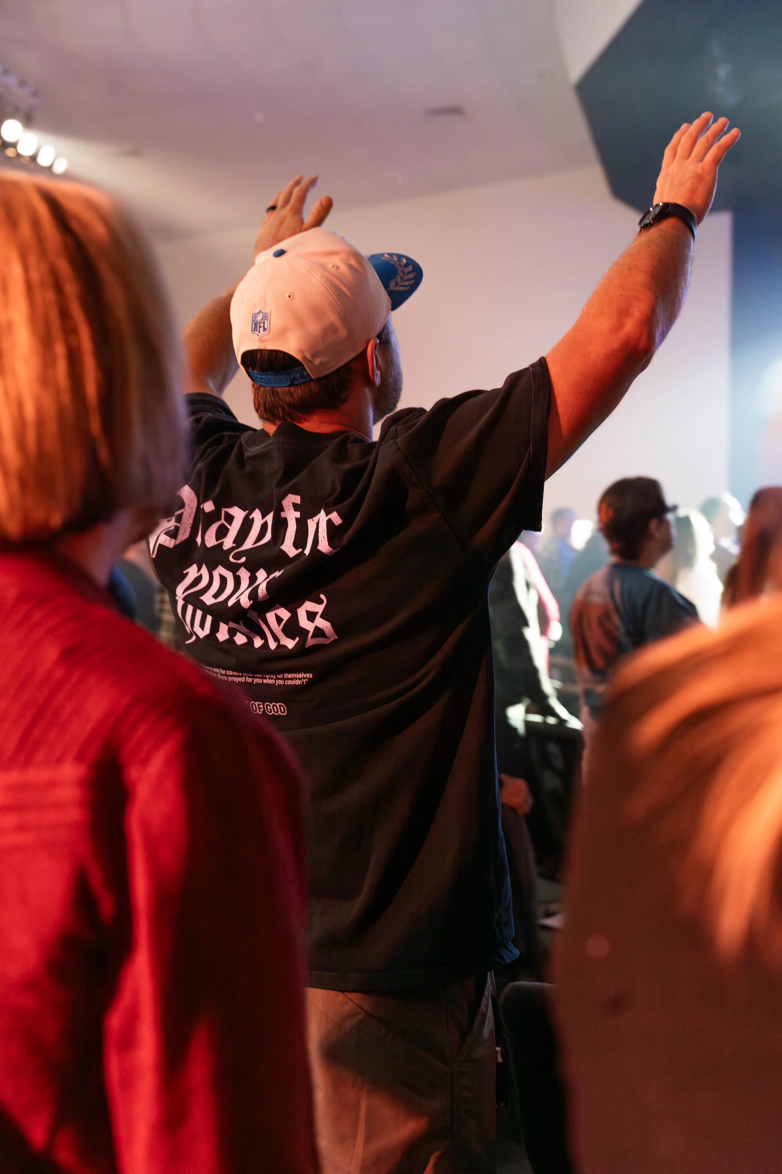 A man wearing a black T-shirt and a white cap with a blue design is raising his arms during a crowd gathering or event in a dimly lit indoor space.
