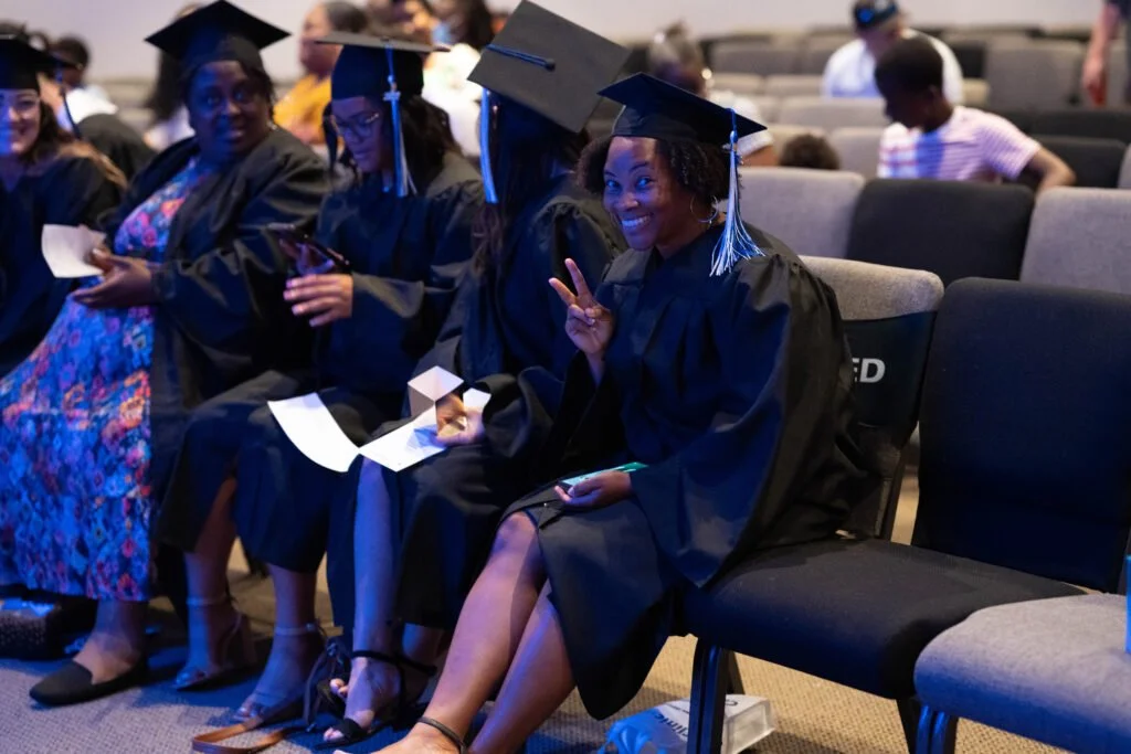 Group of graduates in caps and gowns sitting in an auditorium, with one woman flashing a peace sign and smiling.