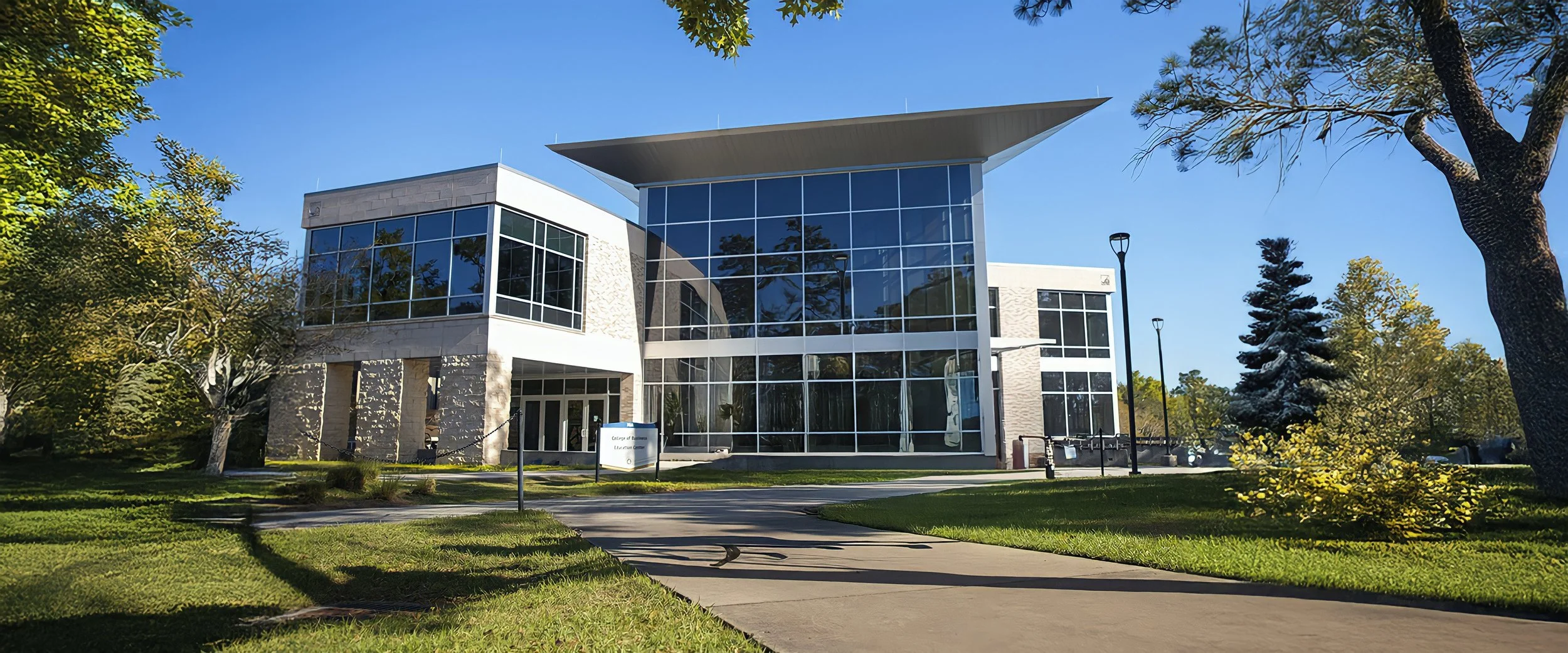 Modern building with large glass windows surrounded by trees, with a pathway leading up to it under a clear blue sky.