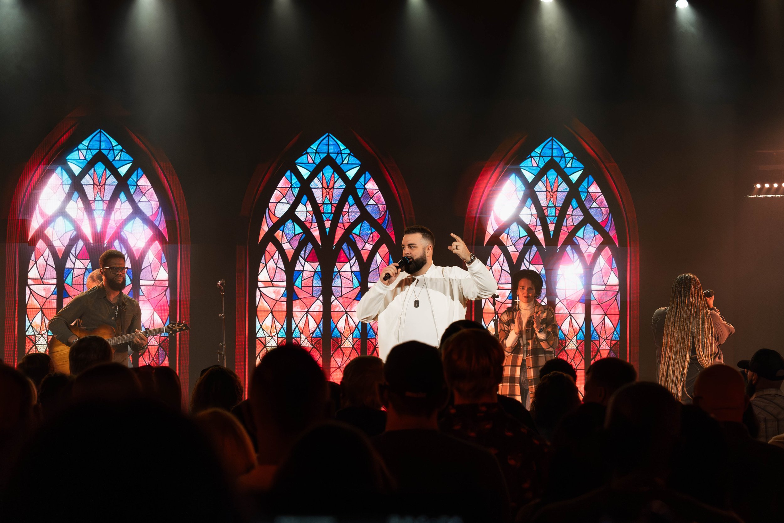 A band performing on a stage with stained glass windows in the background, audience watching.