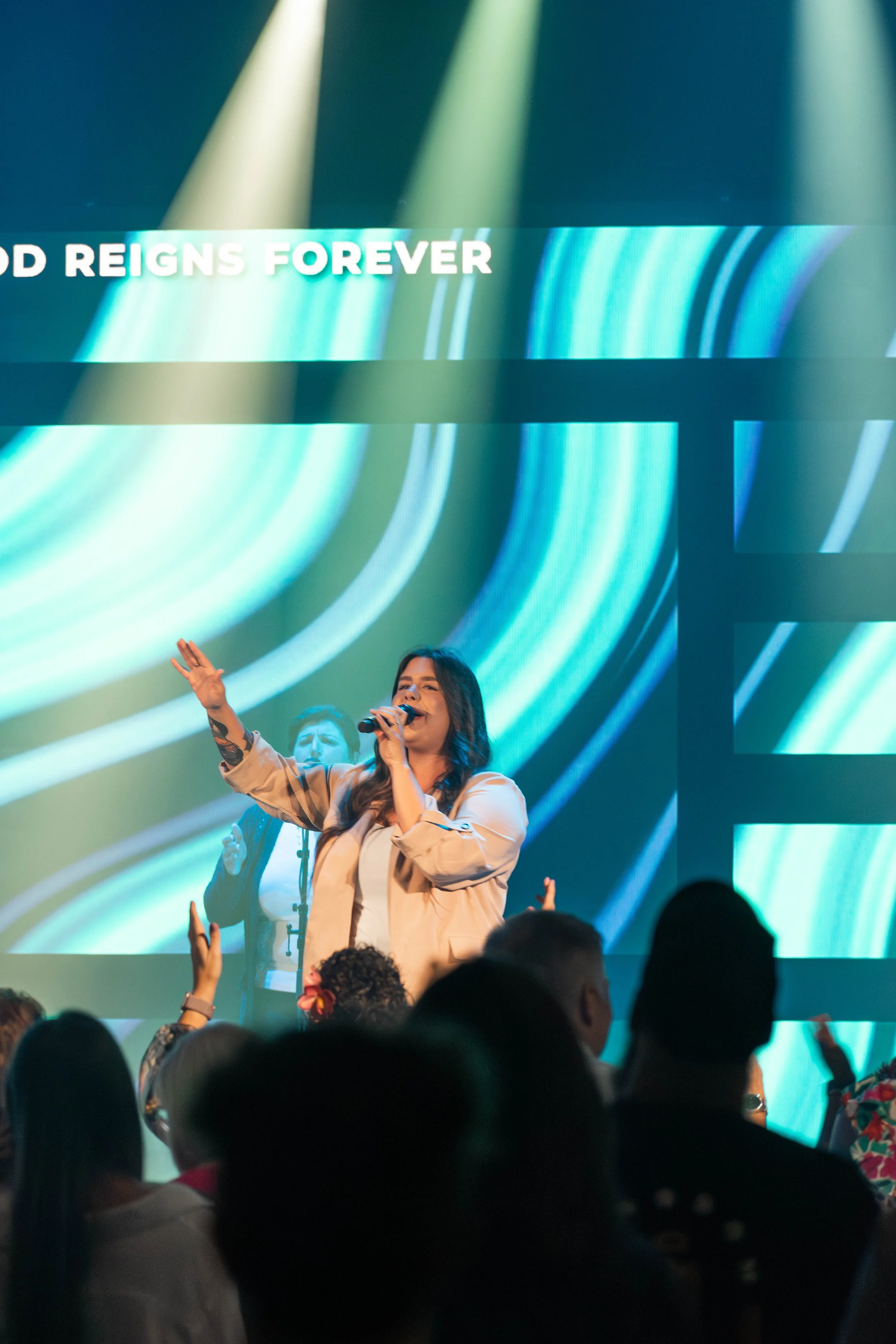 A woman singing on stage with a microphone, audience members with raised hands, colorful background display, and large text reading "God Reigns Forever."