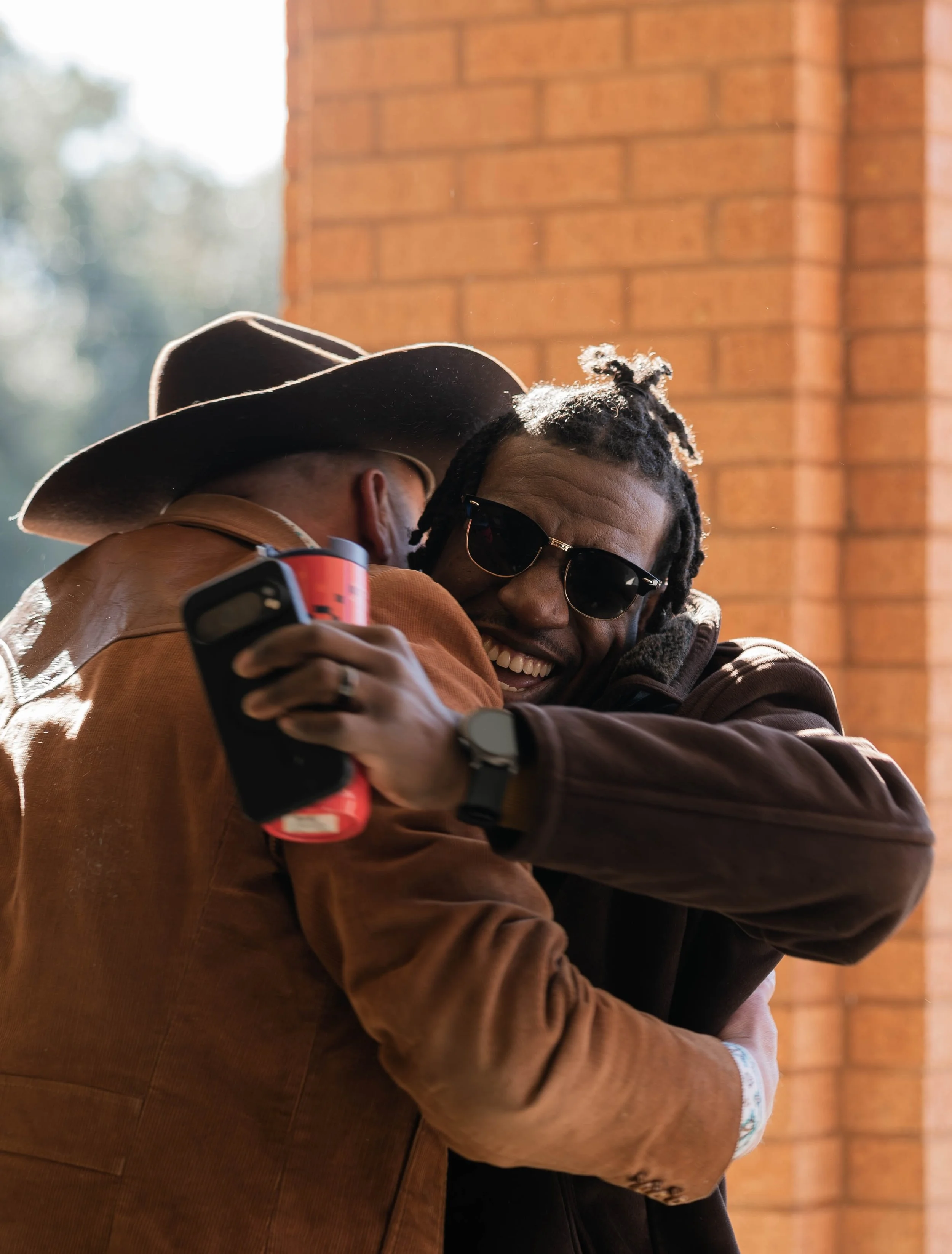 Two men hugging and smiling, one wearing a cowboy hat and sunglasses, the other holding a drink can, outside with brick wall in background.