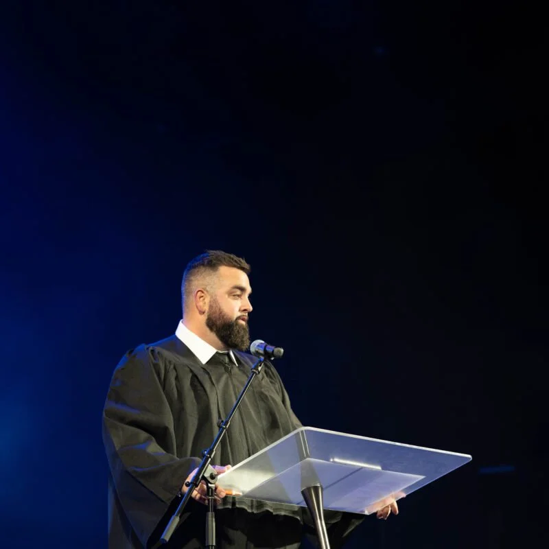 A man in a graduation gown stands at a podium with a microphone, in a dark setting, possibly at a graduation ceremony.