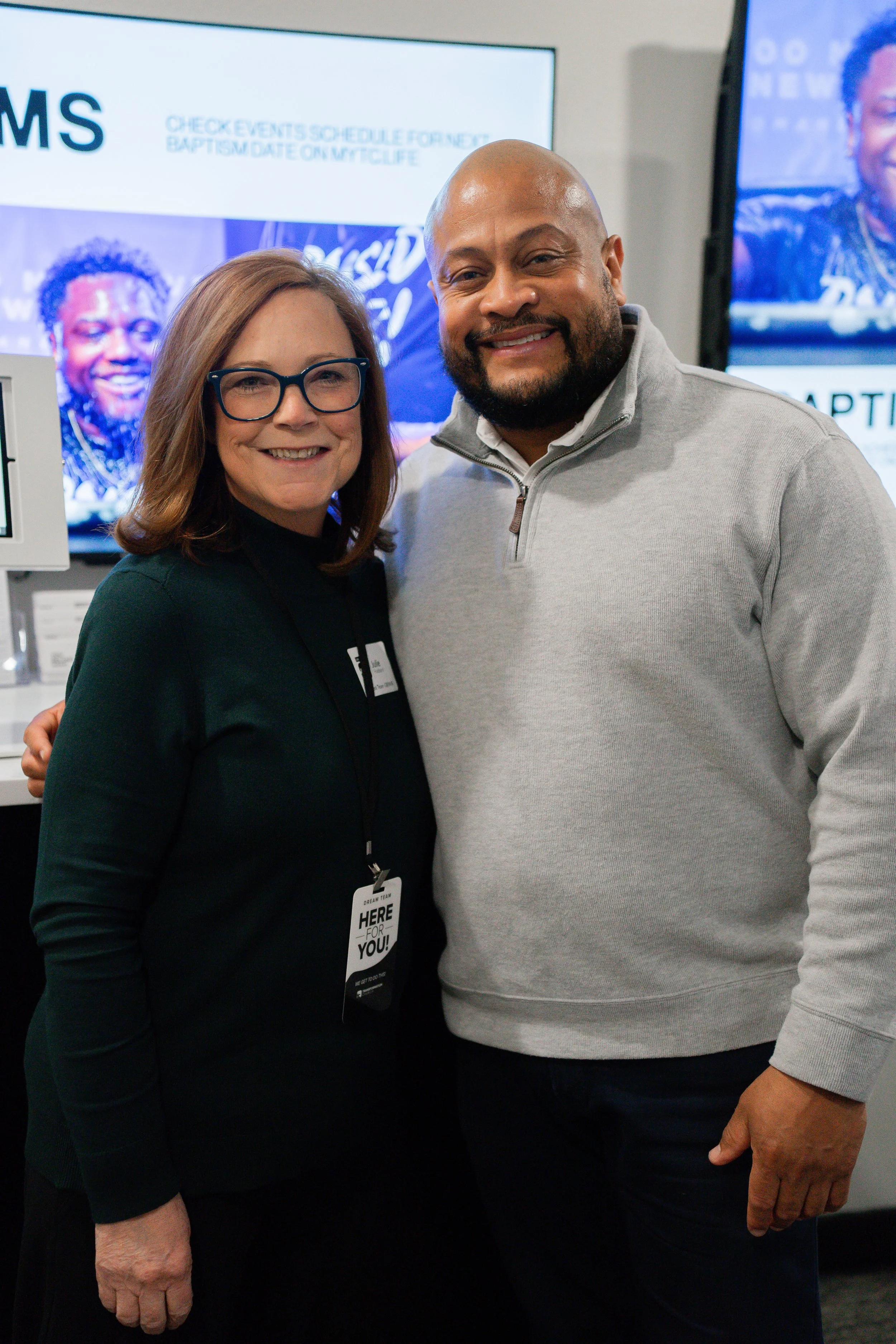 A woman with glasses and a man with a beard smiling and posing together at an indoor event.
