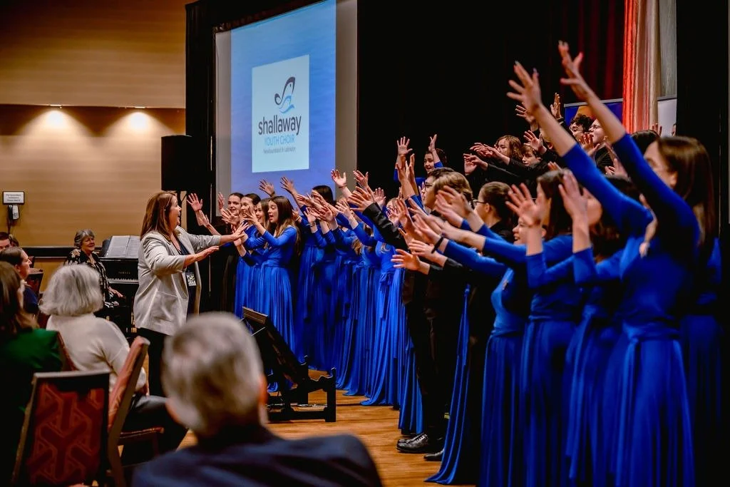 A woman conducts a choir, who are wearing dark blue dresses with their hands raised in the air, while people sit and watch and smile.