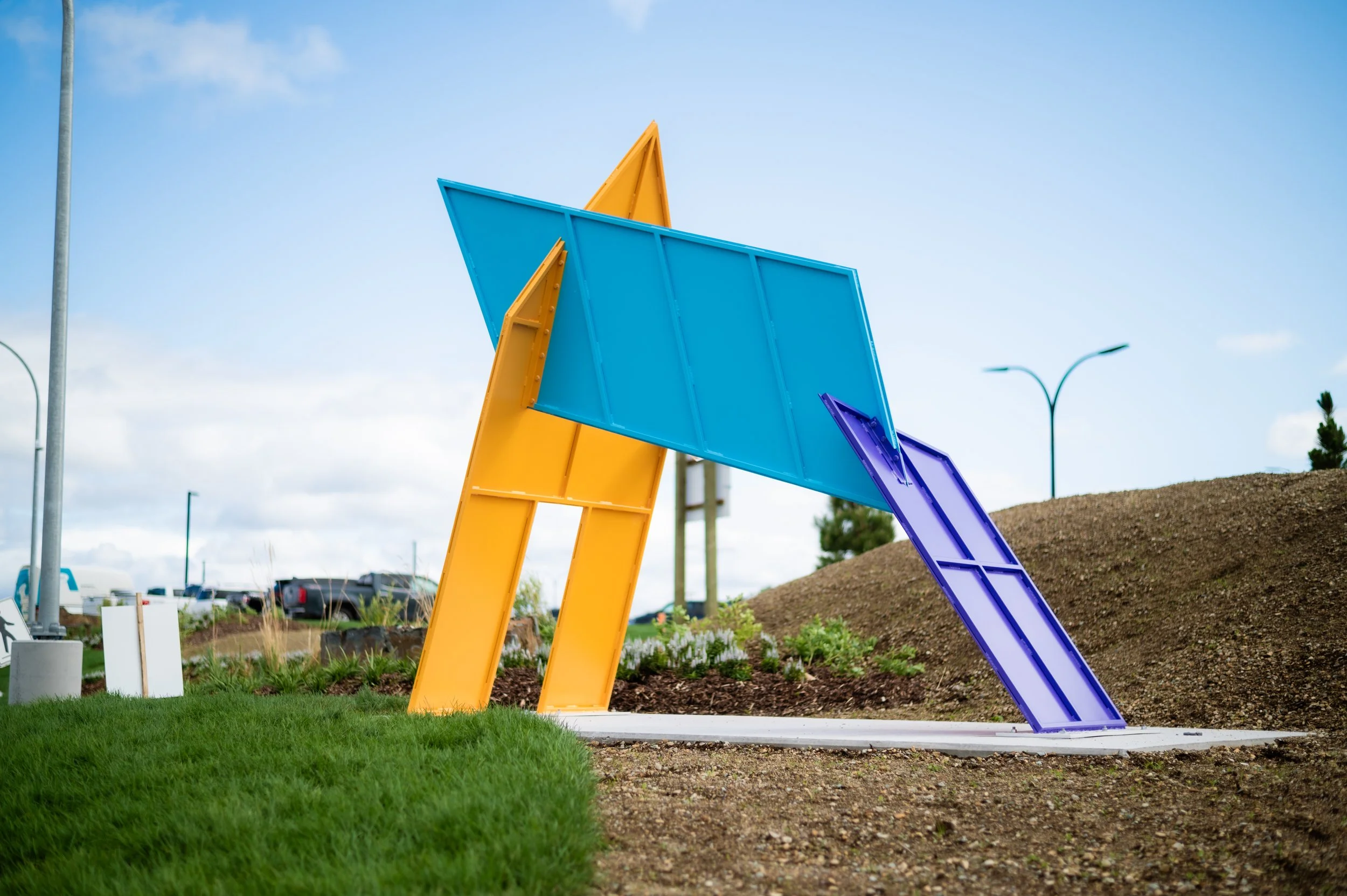 An outdoor sculpture consisting of geometric shapes in yellow, blue and purple lock together. The setting is an airport parking area.