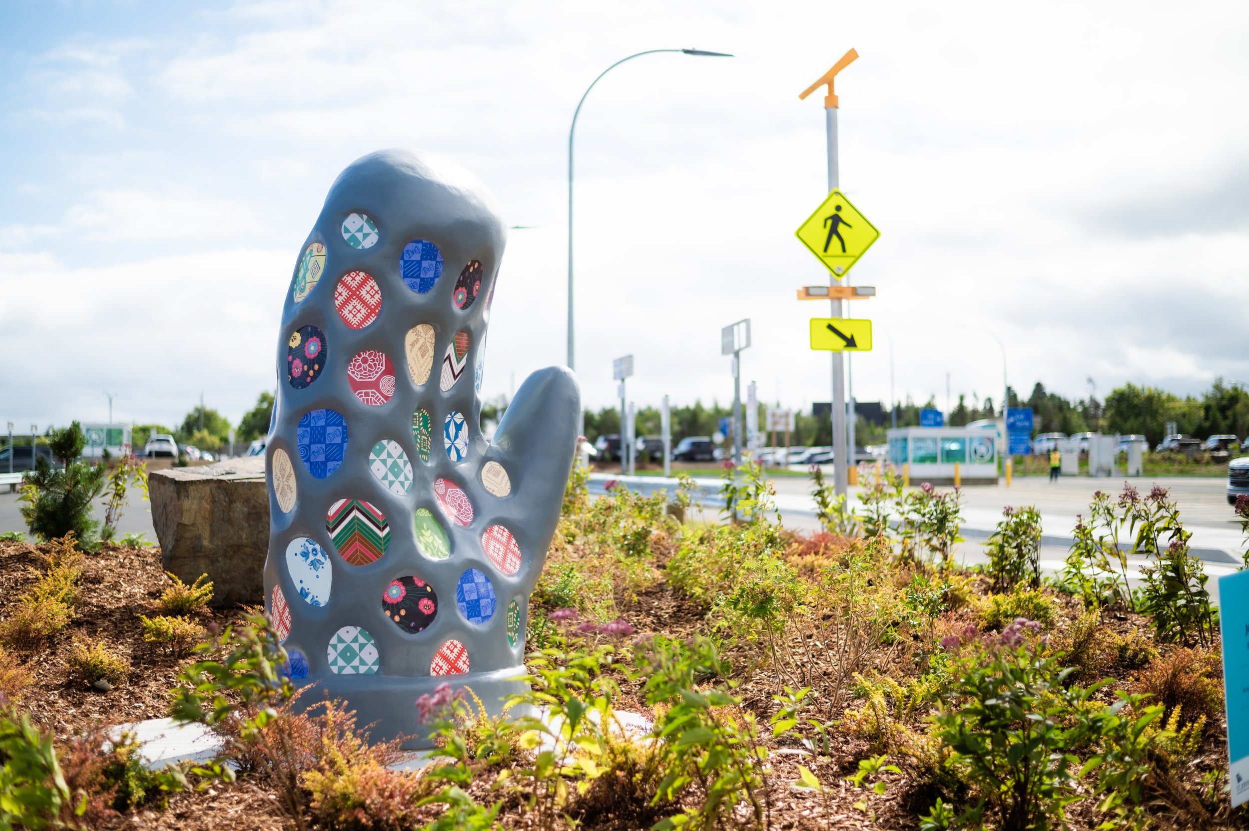 A large sculpture of a mitten with various circular designs embedded within it stands surrounded by small green plants alongside a parking area.