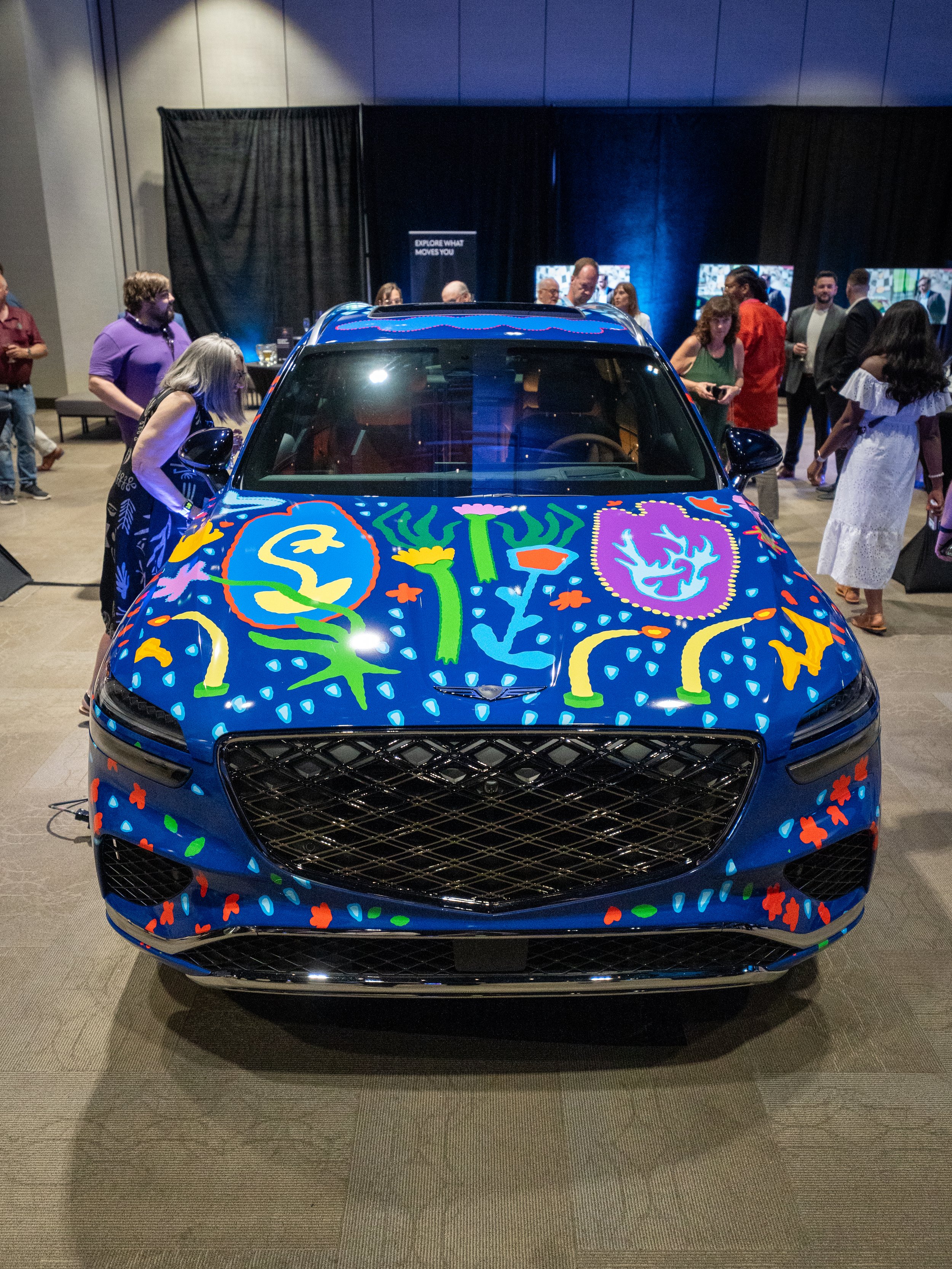 A group of people stand inside an exhibit space and look at a brightly decorated blue car, featuring floral patterns and candles blowing in the wind.