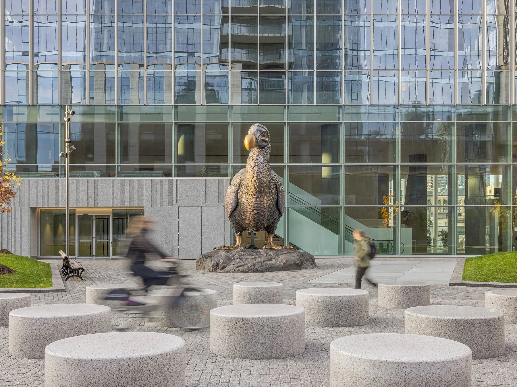 A large statue of a bird sits outside a multi-windowed building as a couple of blurred figures walk and cycle past among concrete cylinders for sitting.