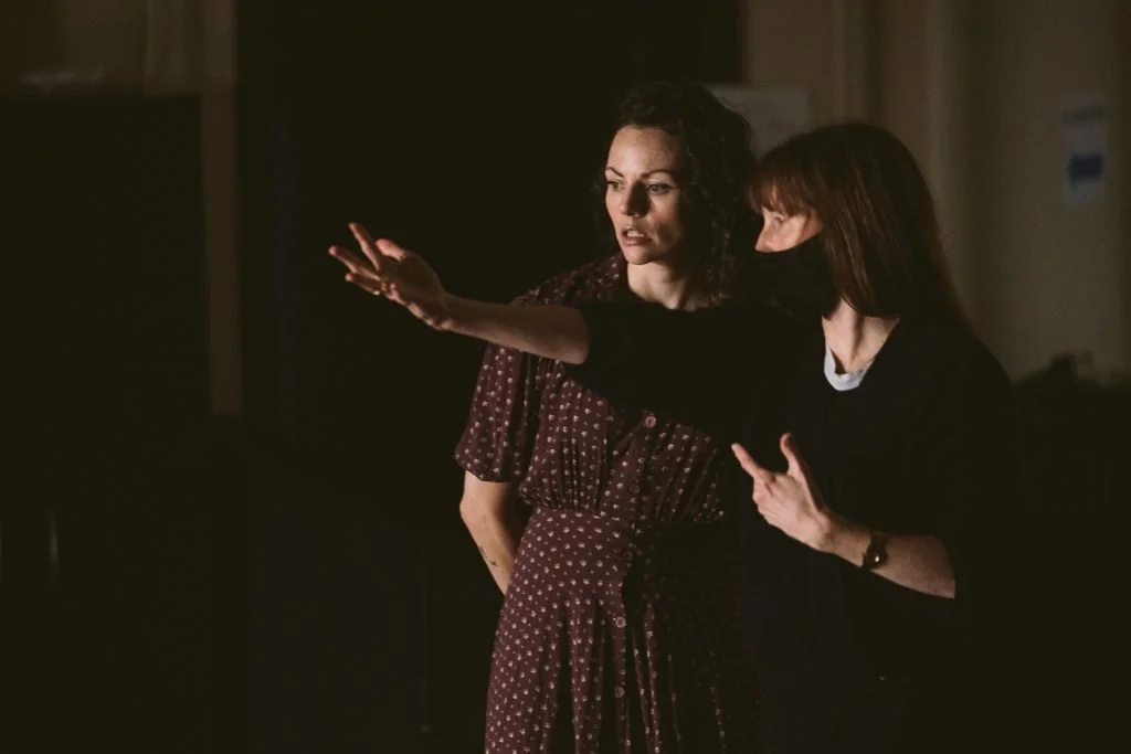 Andrya Duff is wearing a mask and a dark shirt and has her arm outstretched as she provides direction to Bridget Wareham standing next to her. Bridget is wearing a dress and they are both looking at something a bit in the distance.