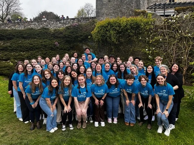 A large group of young people, members of Shallaway Youth Choir, wearing blue t-shirts printed with the choir's name, smile together. Behind them is a stone wall and lush greenery.