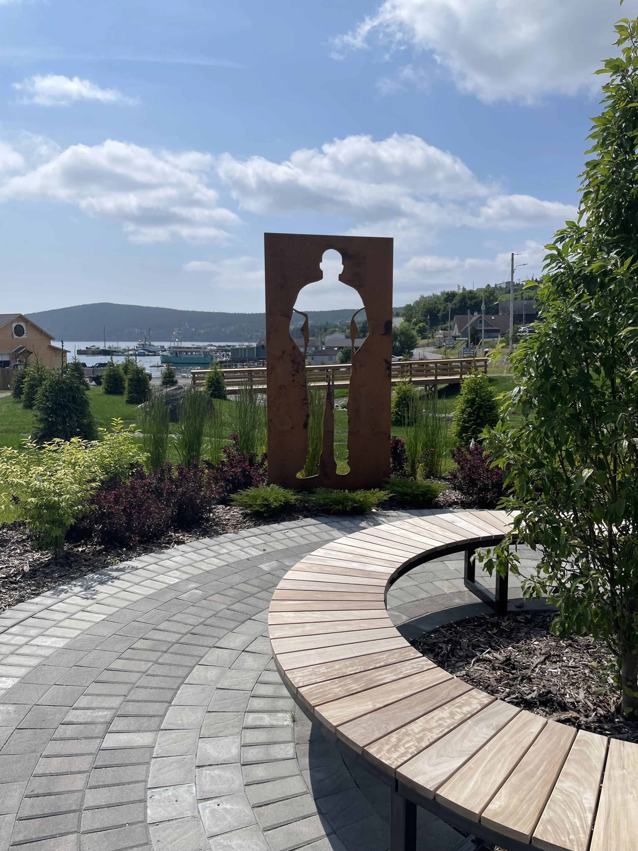 A metal cutout of a person stands in a grassy space,  next to a brick walkway and seating area, with a harbour visible in the background.