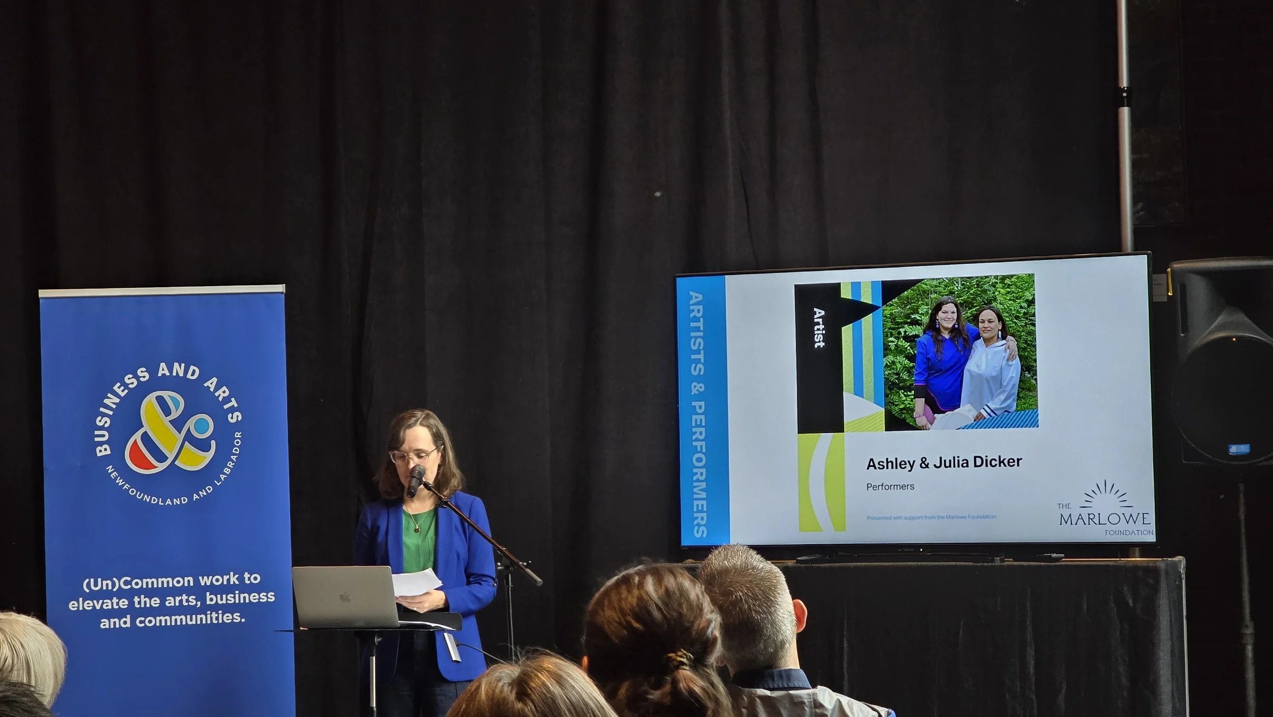 A woman in a blue blazer, green shirt and glasses with shoulder length dark hair speaks into a mic at a podium. Behind her is a blue Business & Arts NL banner and a screen showing two smiling woman, one with her arm around the other.