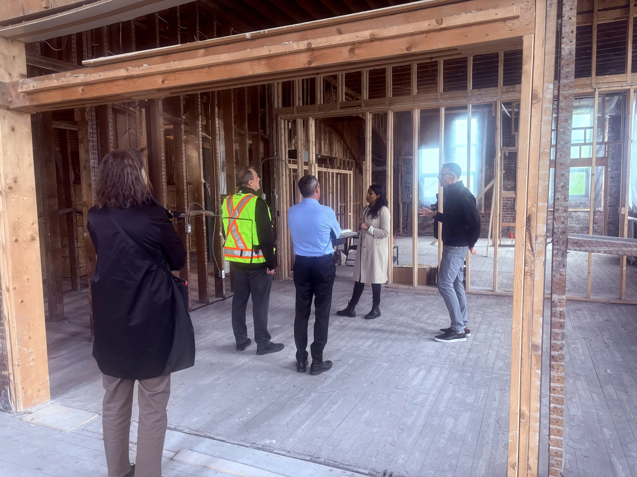 Five people stand and talk while standing inside a room undergoing renovations, with wooden beams exposed. One of the people wears a yellow safety vest.