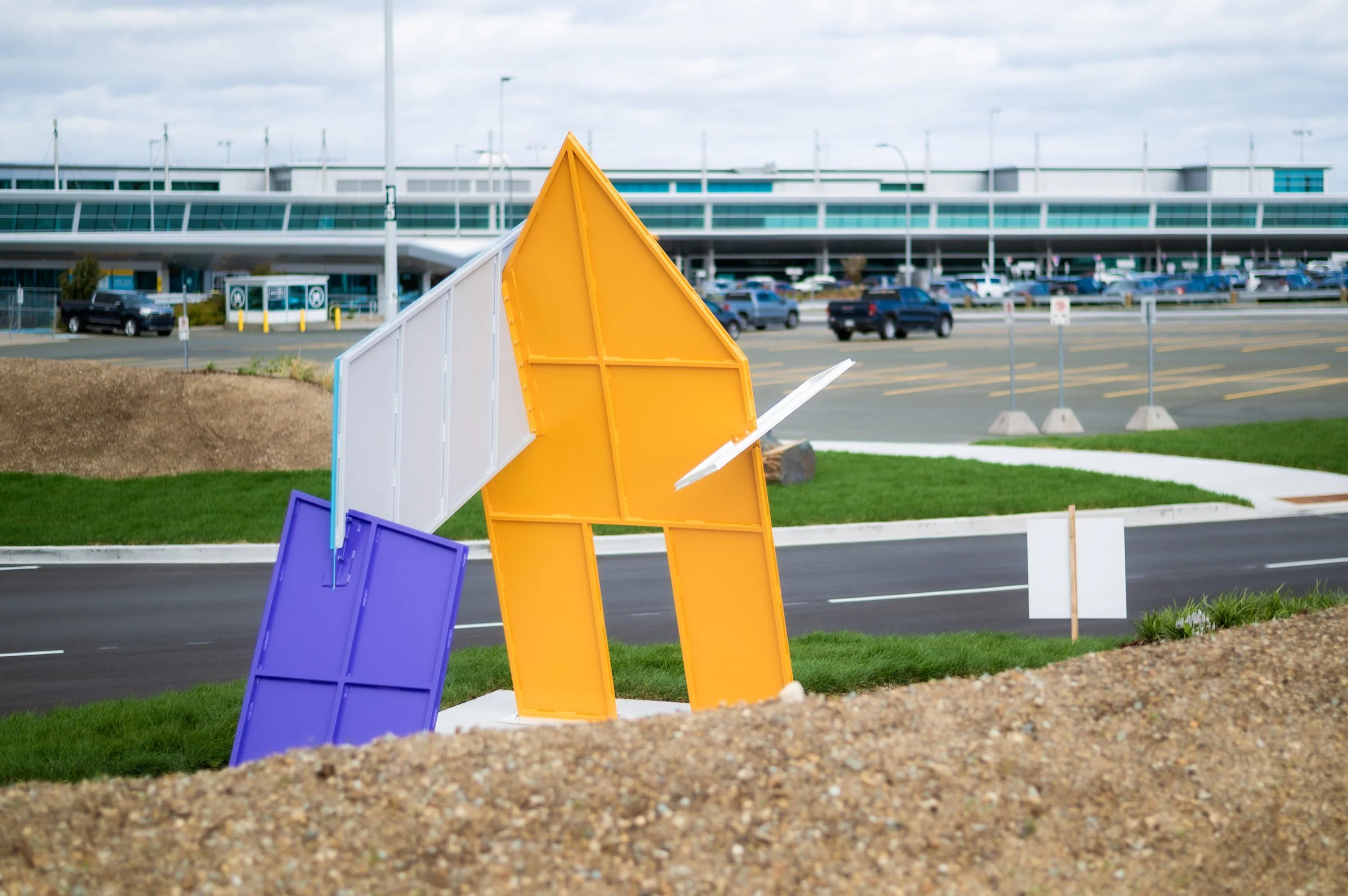 An outdoor sculpture consisting of yellow, white and purple geometric shapes that lock together sits in an airport parking area.