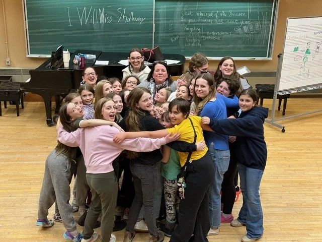 A group of children in a classroom/music room setting, with blackboard in back, smile and laugh as they group hug a woman in the centre.