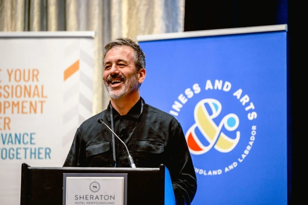 A man with short white/grey hair and a short beard laughs at a podium. Behind him are two banners, including a blue one from Business & Arts NL.
