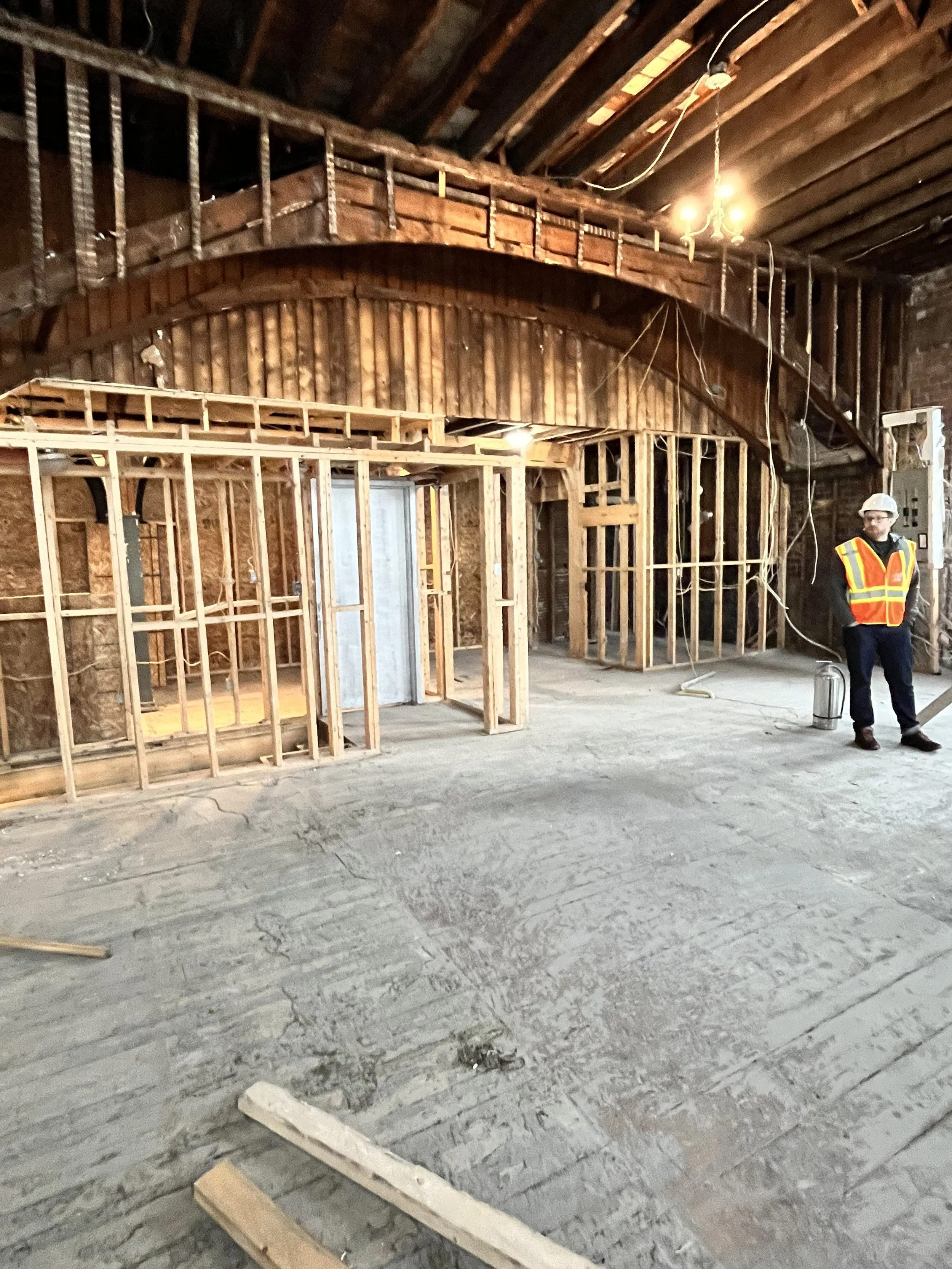 A man in a hard hat and orange vest stands in a room undergoing renovations with wooden walls exposed.