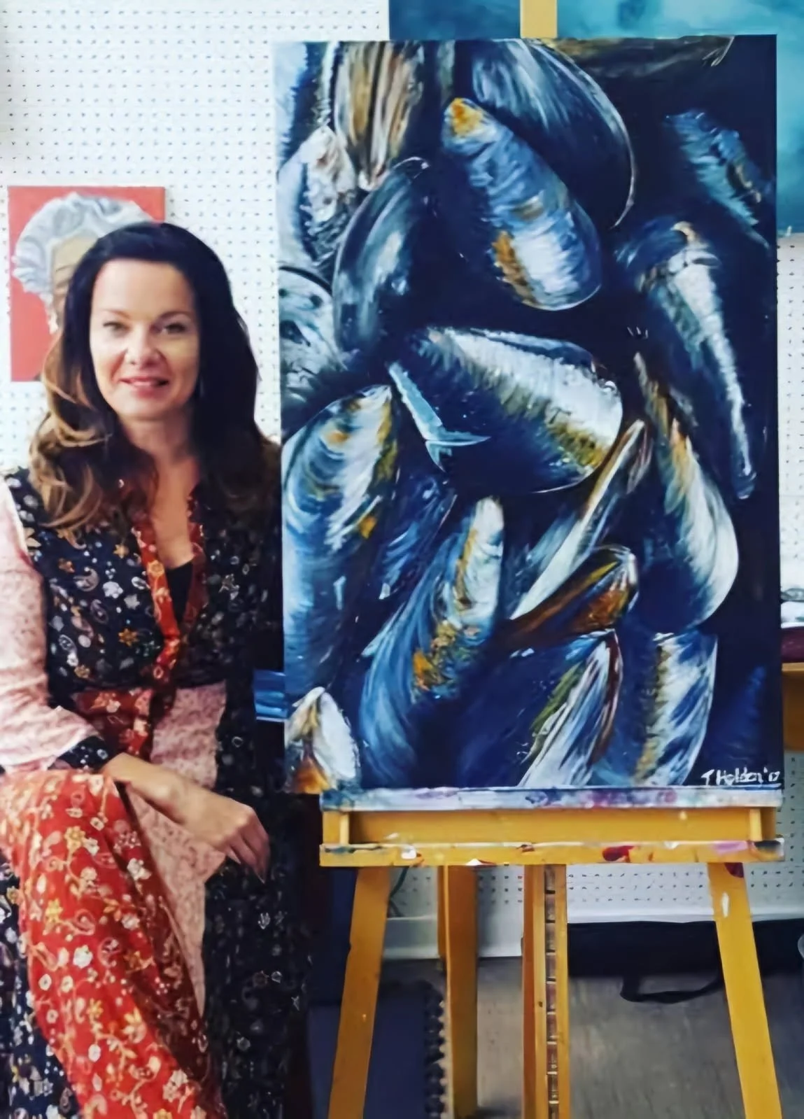 A woman with long brown hair, wearing a long dress in a blue and red floral pattern, sits and smiles next to an easel holding a painting of mussel shells.