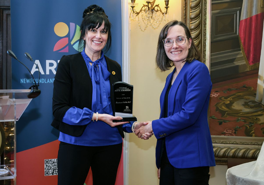 Two woman, the one on the left wearing a blue collard blouse and black jacket, and the one on the right wearing a blue blazer, both smile at the camera as they shake hands, with the woman on the left handing the one on the right an award.