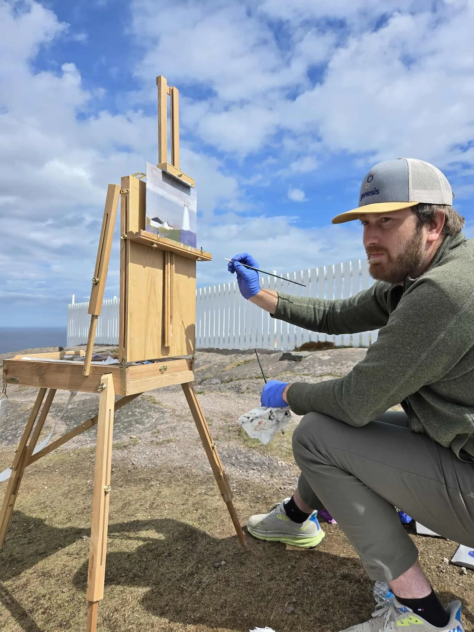 A mean in a ball cap crouches on a rocky landscape and looks to the left as he raised a gloved hand to an easel, upon which sits a painting of a lighthouse. A long white fence runs along his left side and ahead is the ocean.