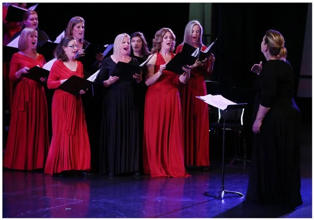 A woman standing at a podium in a long black gown directs members of a choir who are wearing red gowns and holding sheet music in a book.