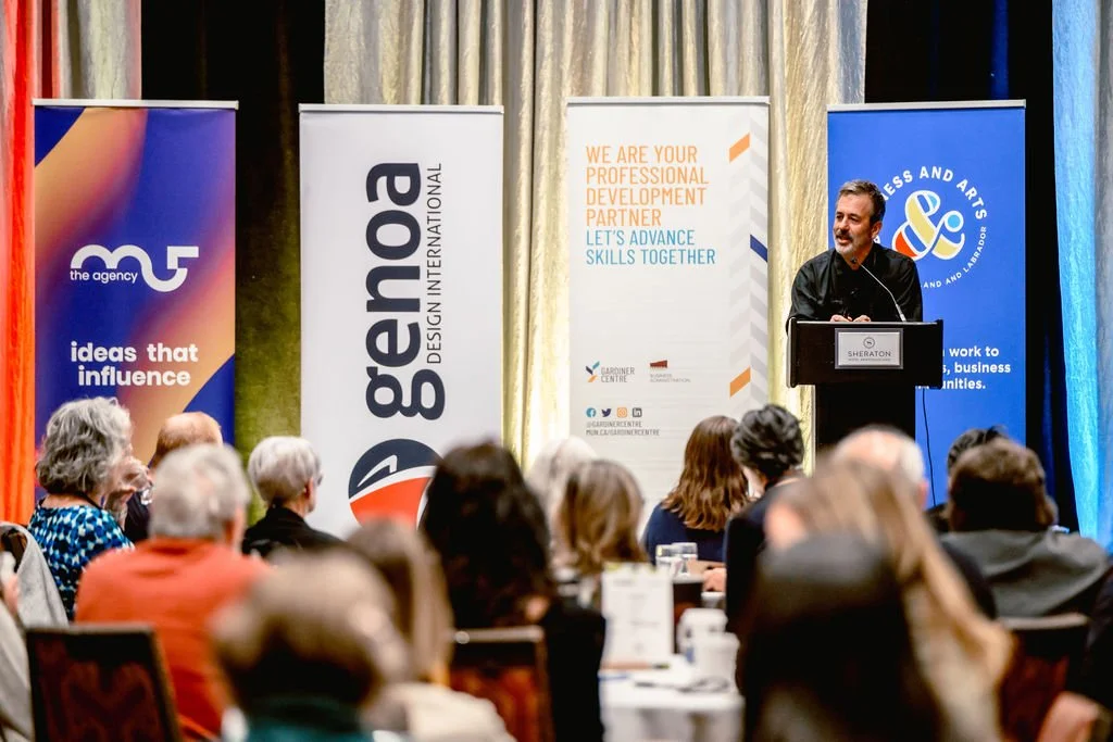A man in a dark shirt speaks from a podium to a seated crowd. Behind him are stand-up banners from M5 the Agency, Genoa Design International, Gardiner Centre and Business & Arts NL.