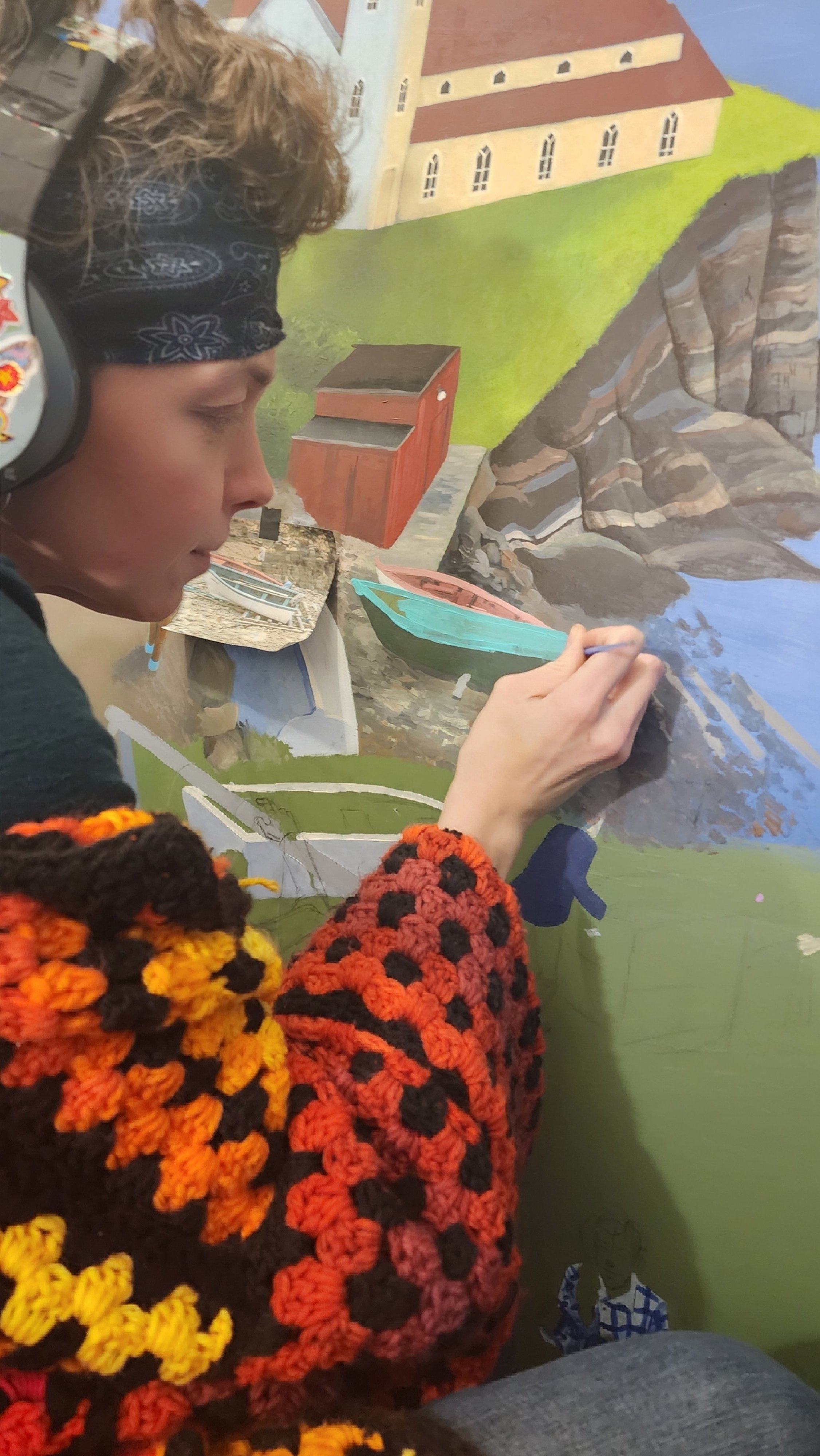 A close-up shot of an artist in a bright crocheted sweater, painting a section of a boat on a mural from a photo tacked alongside her.