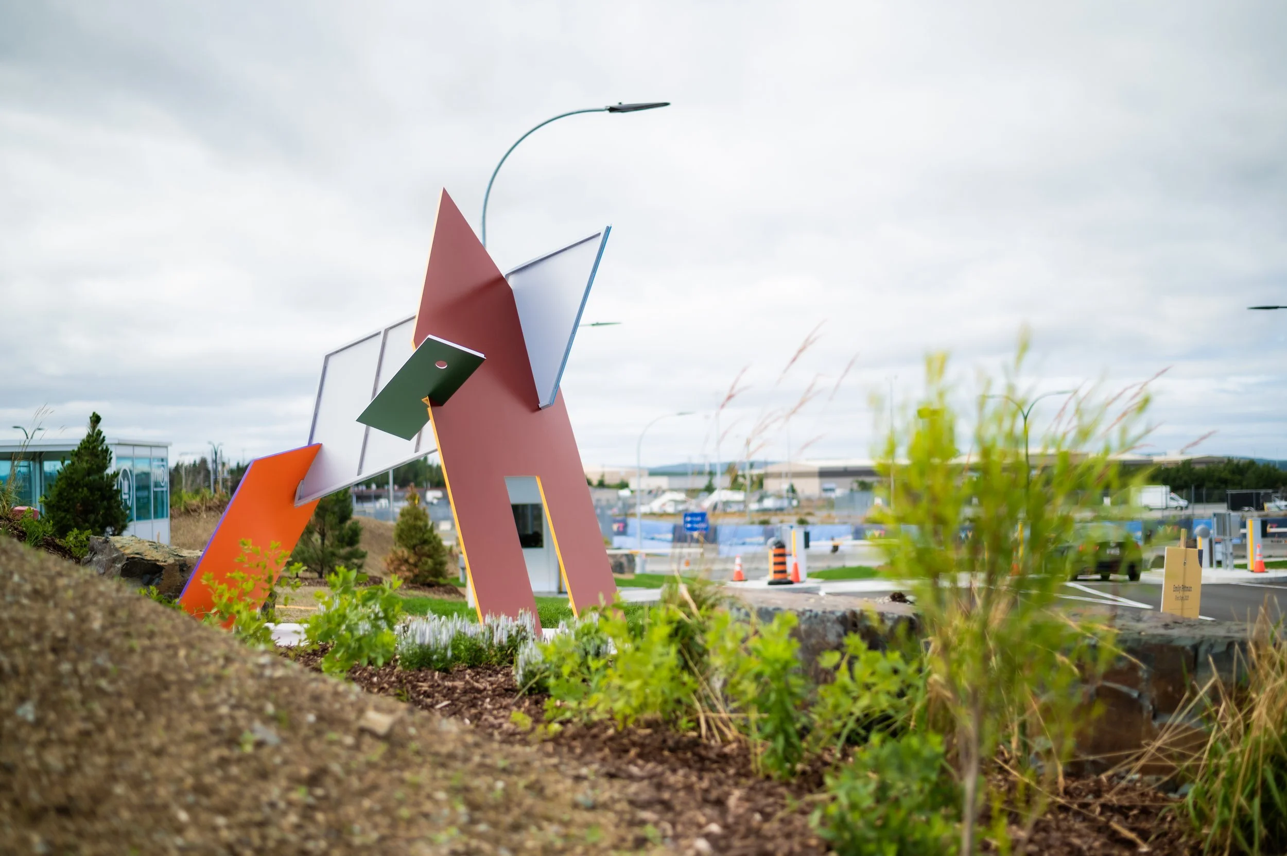 A sculpture consisting of red, orange and white shapes that lock together sits alongside an airport parking area.
