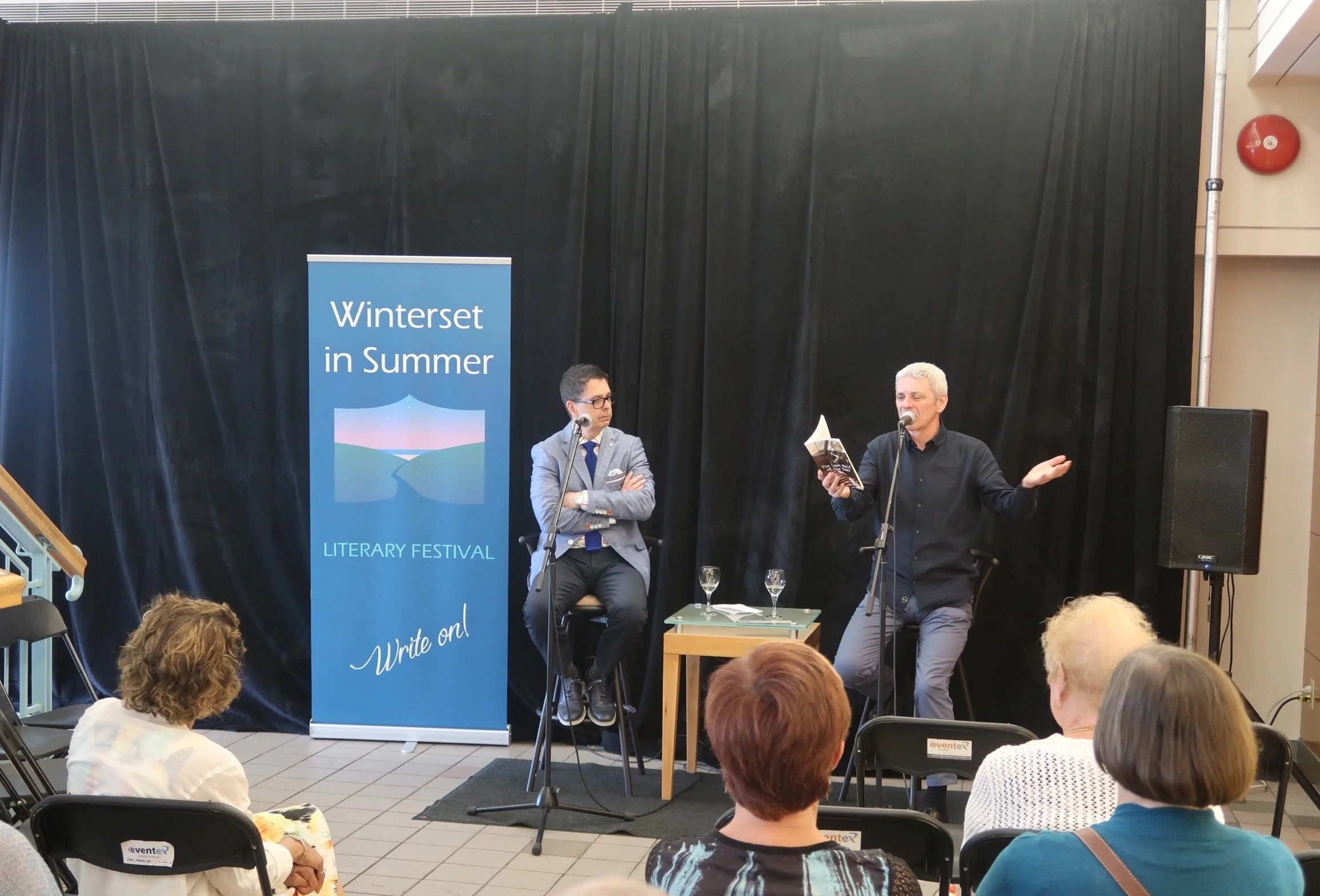 Two men sit in front of a black curtain in front of a seated group. The man to the left watches as the man to the right holds a book and speaks into a microphone. To their left is a banner promoting Winter in Summer Literary Festival.