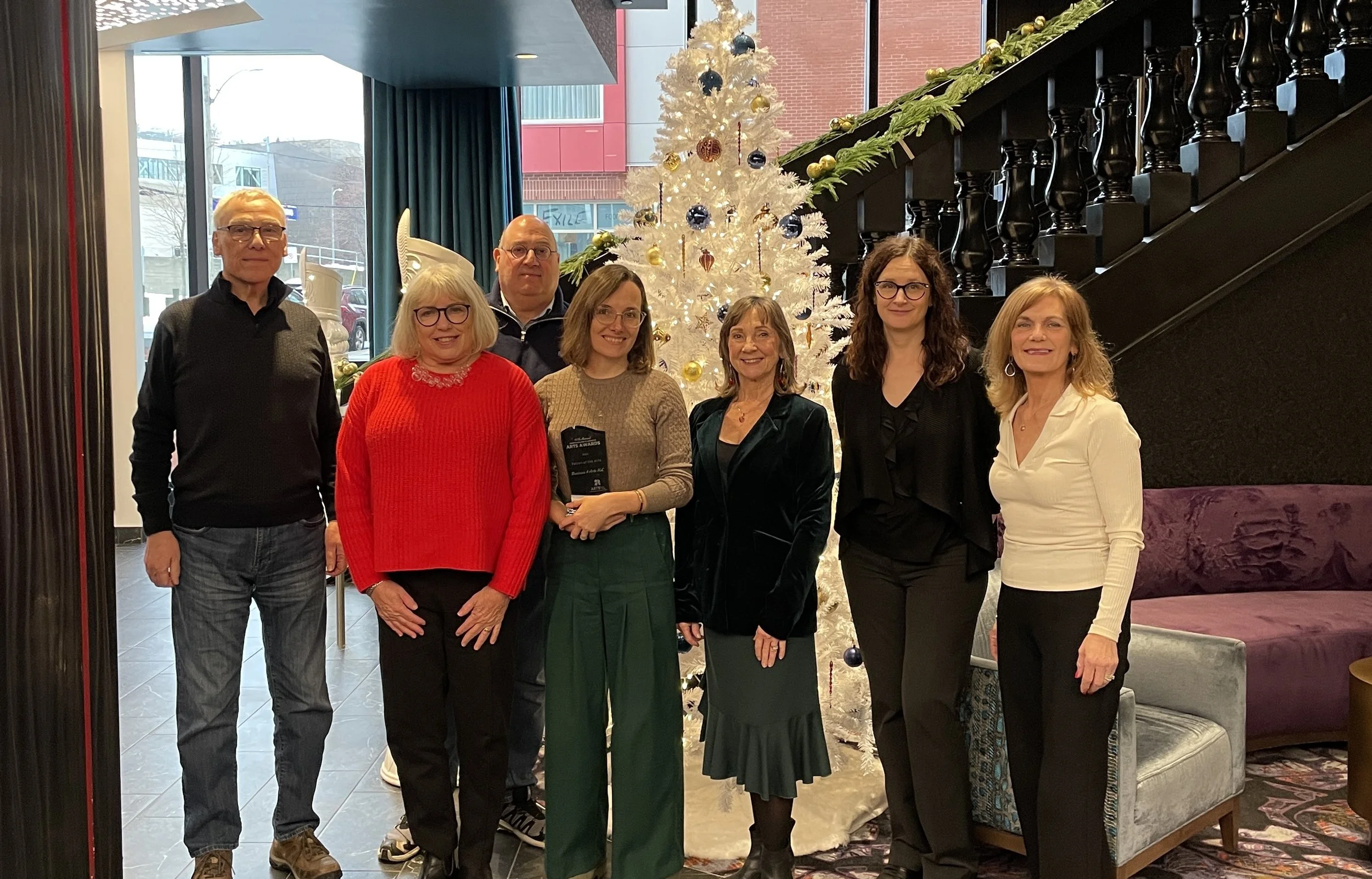 Two men and five women stand together and smile in front of a white Christmas chair with a black staircase in the background. Also behind them are large windows, through which buildings can be seen.